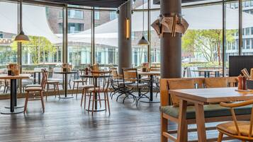 An indoor dining area with wooden tables and chairs, large windows, and hanging pendant lights.