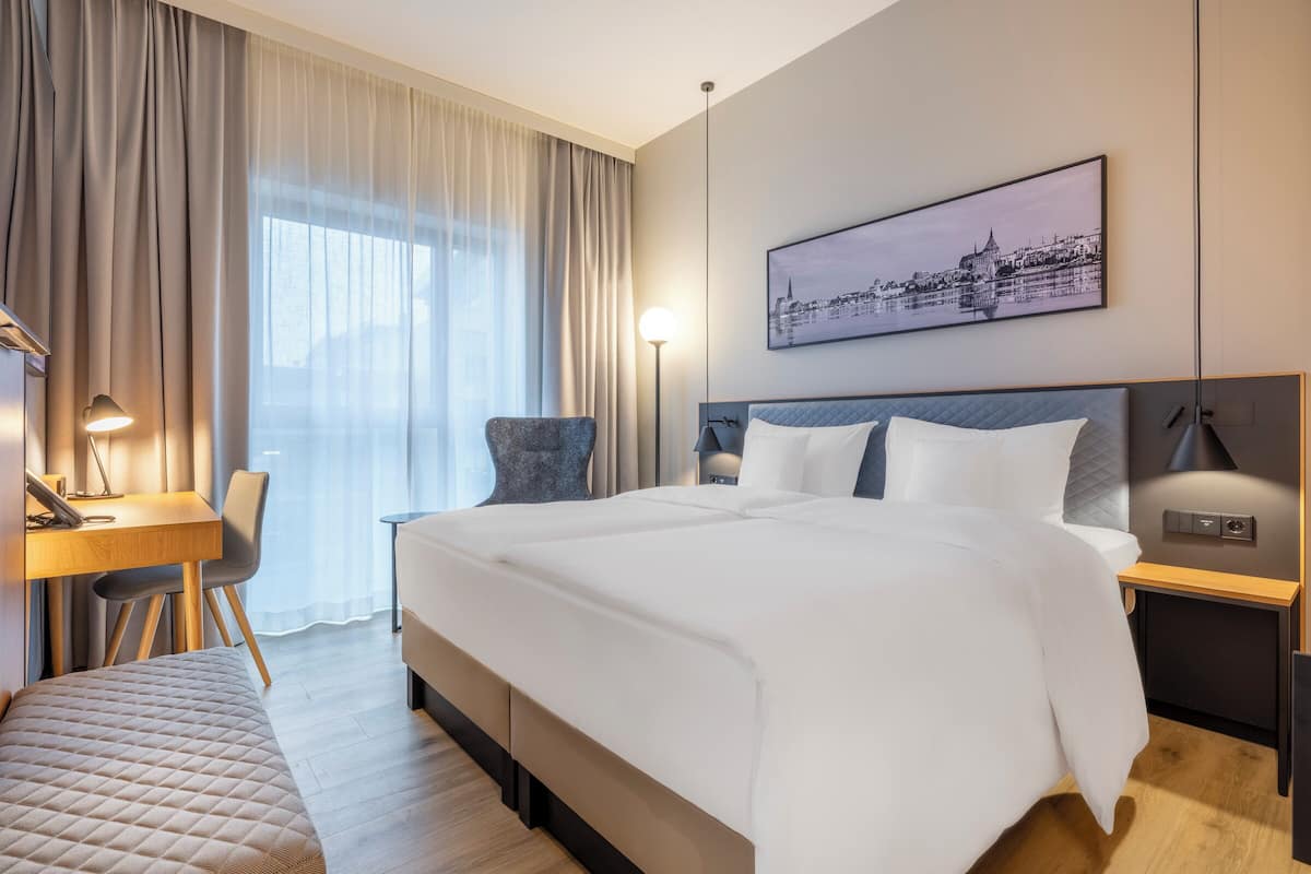 A modern hotel room with a large bed, a desk, a chair, and a view of a cityscape through the window.