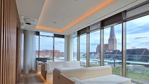 A modern hotel room with a large window, a bed, a seating area, and a view of a cityscape. - Scanhotels Stadthafen - Neueröffnung (Rostock)