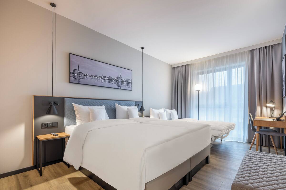 A modern hotel room with a large bed, bedside tables, a desk, and a view of a cityscape through the window.