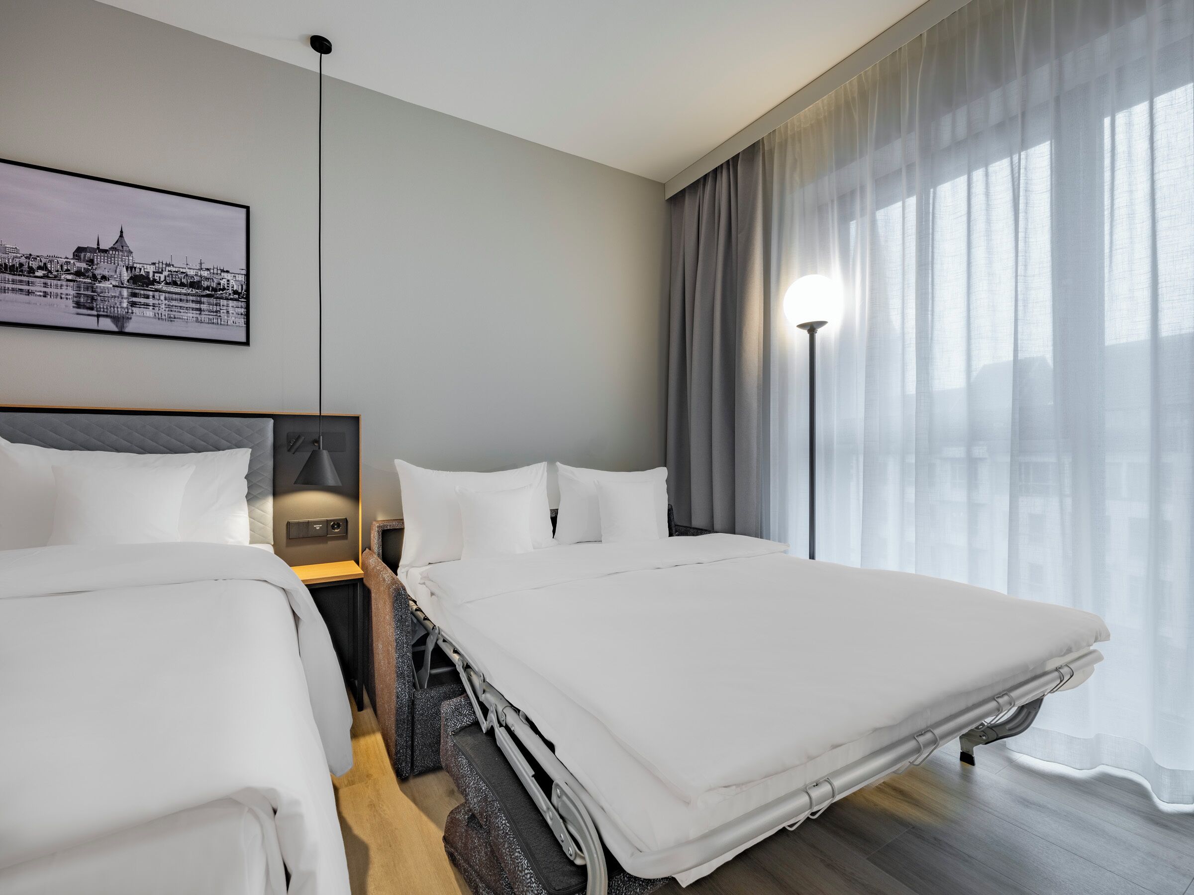 A hotel room with two beds, a cityscape painting, a lamp, and a view of the city through sheer curtains.