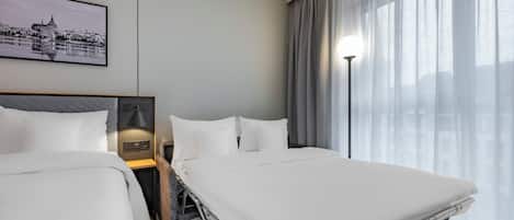 A hotel room with two beds, a cityscape painting, a lamp, and a view of the city through sheer curtains.