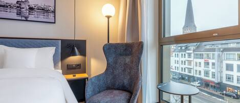 A hotel room with a bed, a chair, a small table, a lamp, and a large window with a city view.