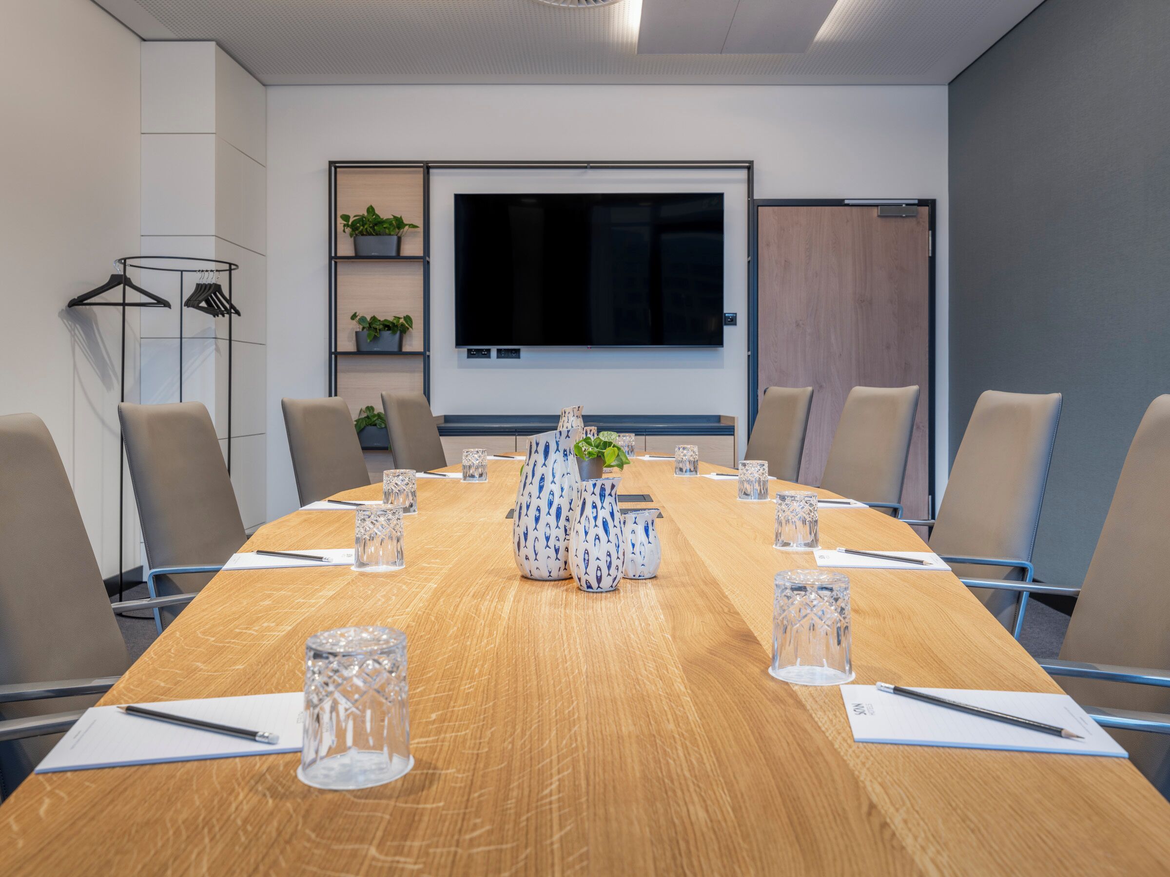 Een vergaderzaal met een lange tafel, stoelen, een televisiescherm en potplanten.