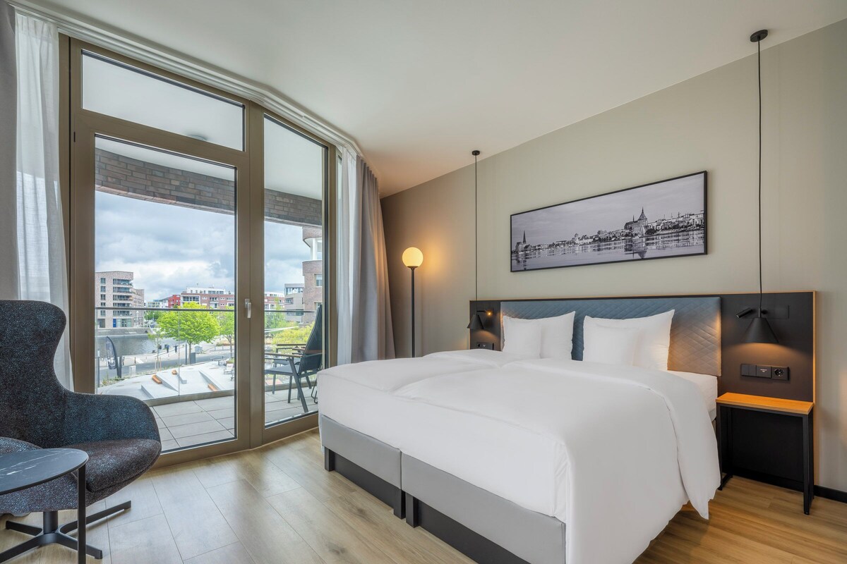 A modern hotel room with a large bed, a desk, a chair, and a cityscape view through the window.