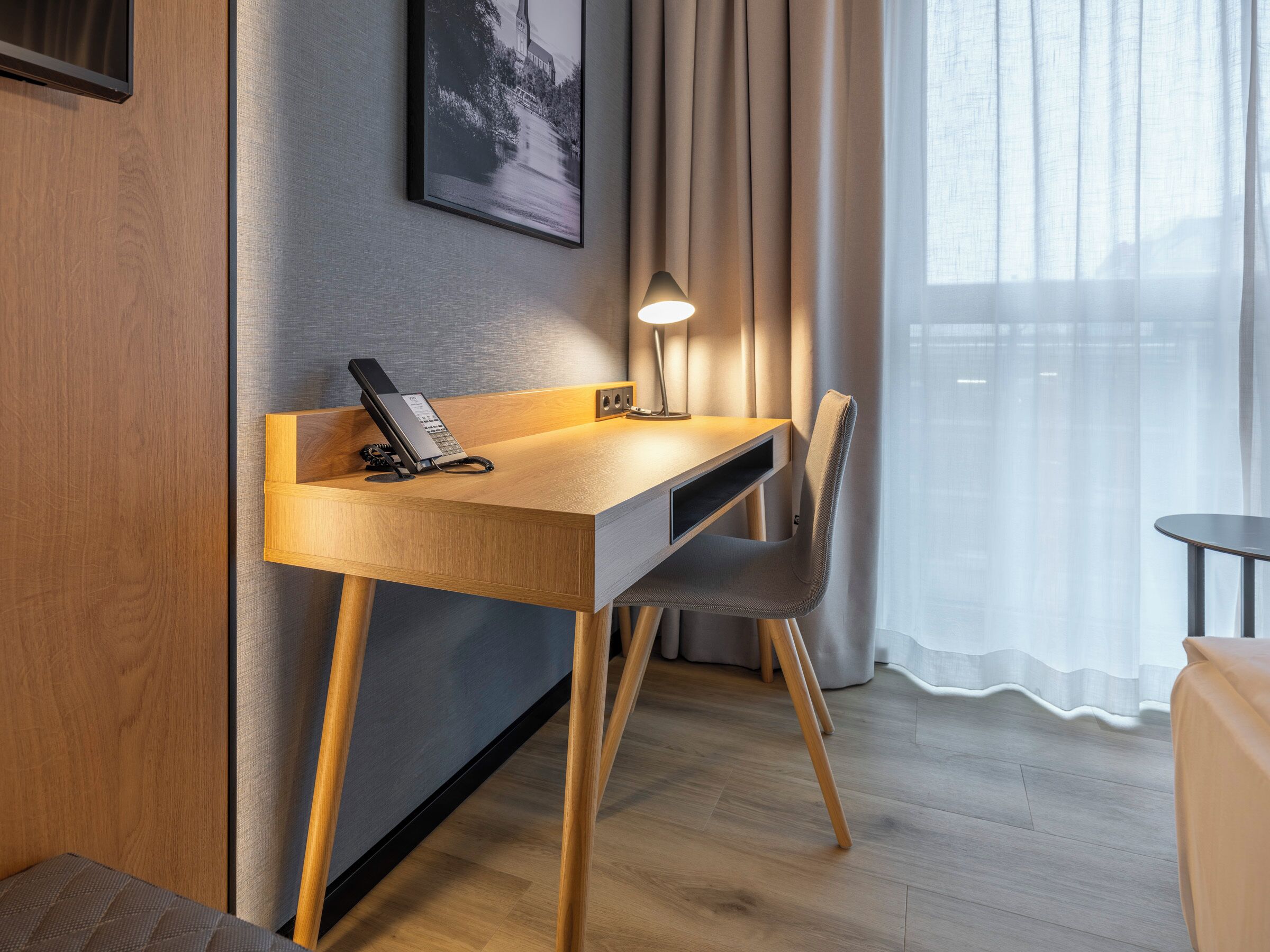 Ein modernes Hotelzimmer mit Schreibtisch aus Holz, einem Sessel, einem Telefon und einer Lampe.