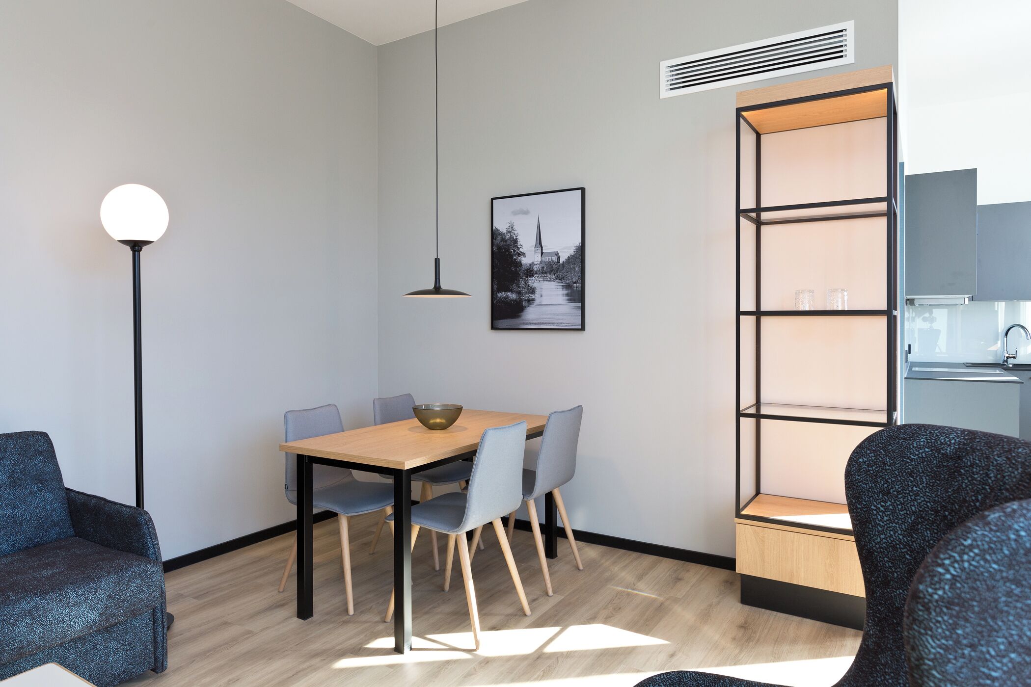 A modern room with a dining table, chairs, a pendant light, a framed picture, a built-in shelf, and a kitchenette.