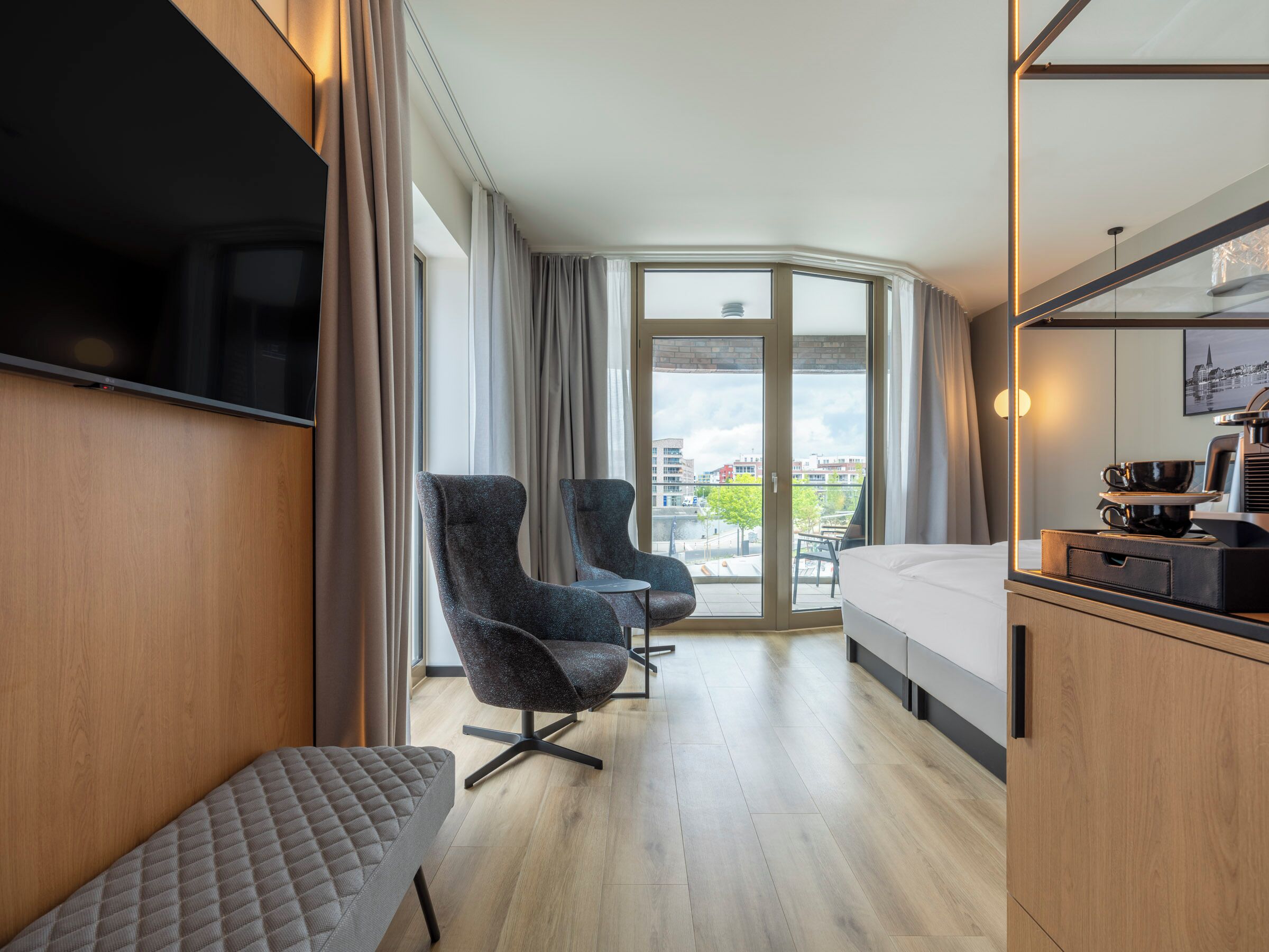A hotel room with a bed, two armchairs, a small table, a TV, and a balcony with a city view.