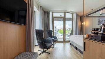 A hotel room with a bed, two armchairs, a small table, a TV, and a balcony with a city view.