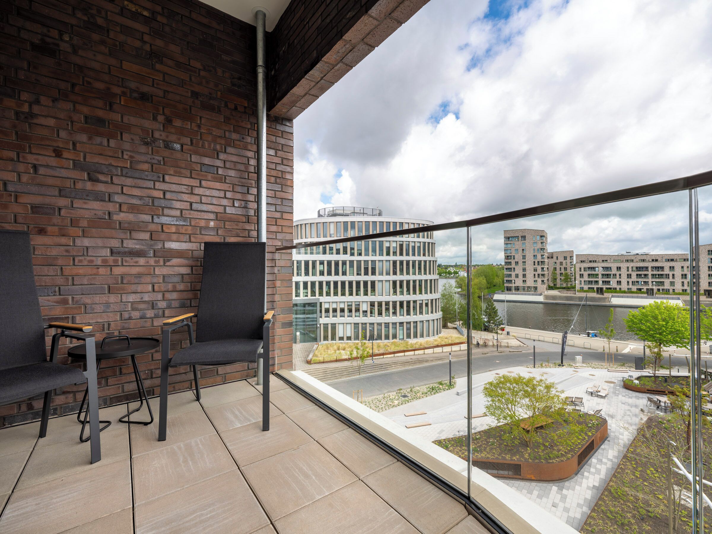 A balcony with a brick wall, two chairs, a table, and a view of a modern building and a river.