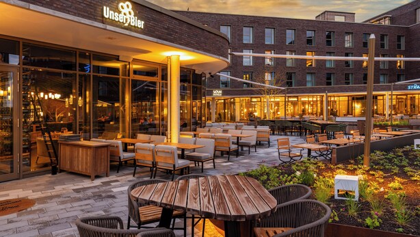 An outdoor seating area with tables and chairs, a brick building with a sign that reads 'UnserBier,' and a clear sky. - Scanhotels Stadthafen - Neueröffnung (Rostock)