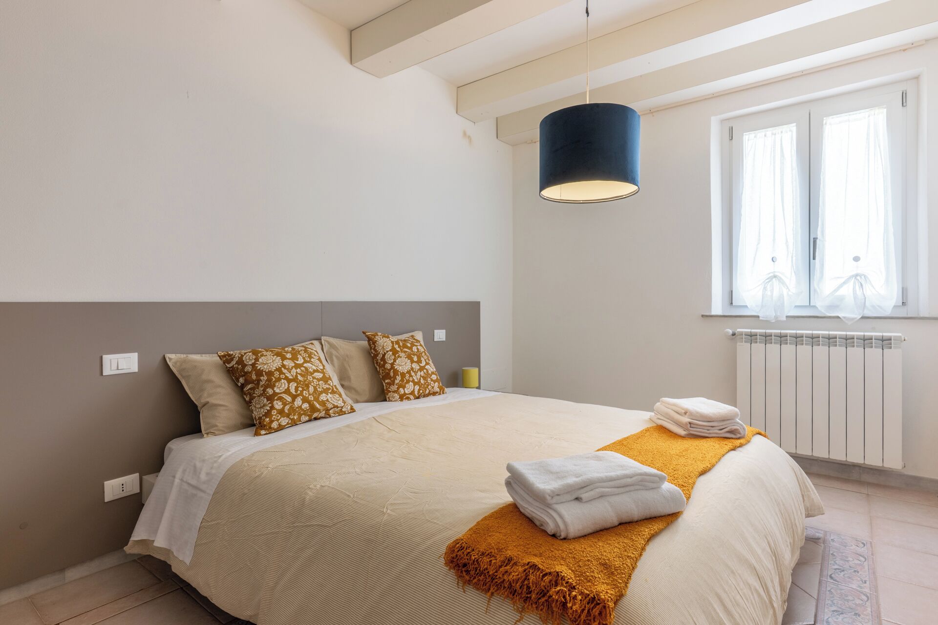 A bedroom with a bed, a bedside lamp, a window with curtains, and a radiator.