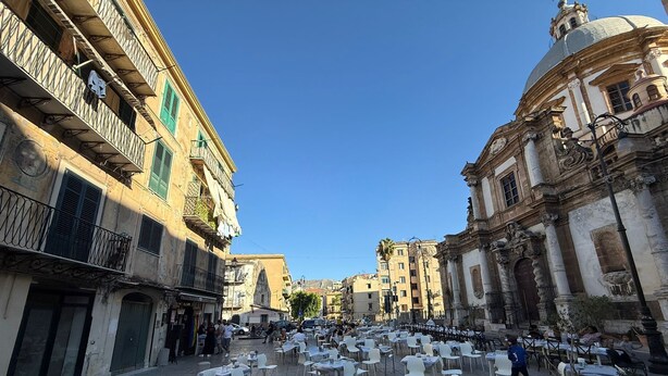 An outdoor café area with tables and chairs, surrounded by historic buildings with balconies and decorative elements. - ABT L'Aura  (Palermo)