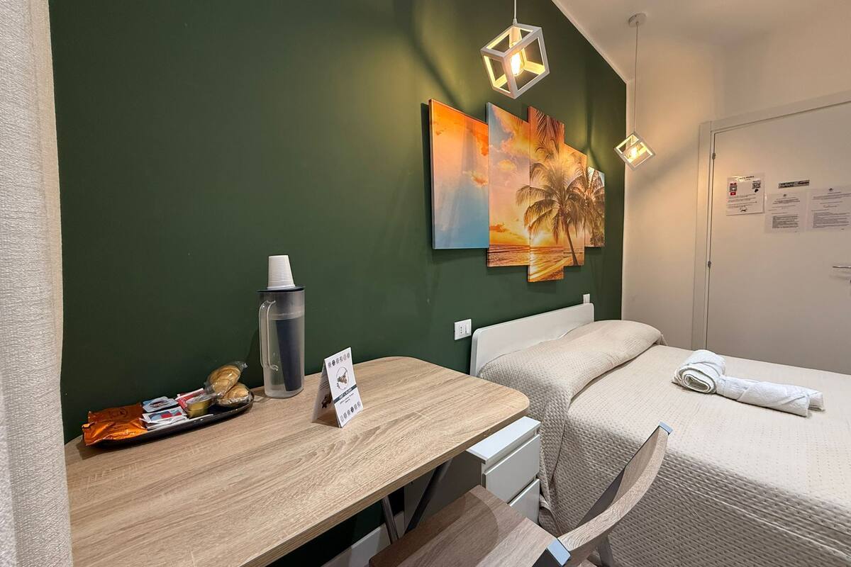 A hotel room with a wooden desk, a bed with a white comforter, and two wall-mounted paintings depicting a sunset and a palm tree.