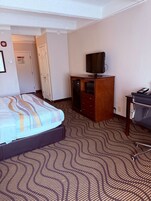 Una habitación de hotel con una cama, un televisor sobre un mueble de madera, un escritorio con una silla y una alfombra estampada.