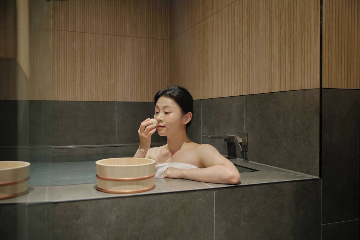 A person in a modern bathroom with a wooden panel wall, a large mirror, and a towel wrapped around their body.