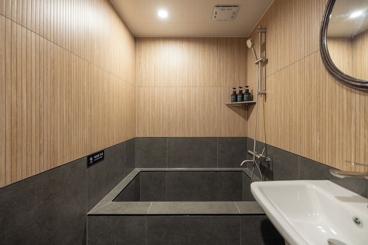 A modern bathroom with a bathtub, a sink, and a mirror. There is a shelf with toiletries and a sign indicating the room type.
