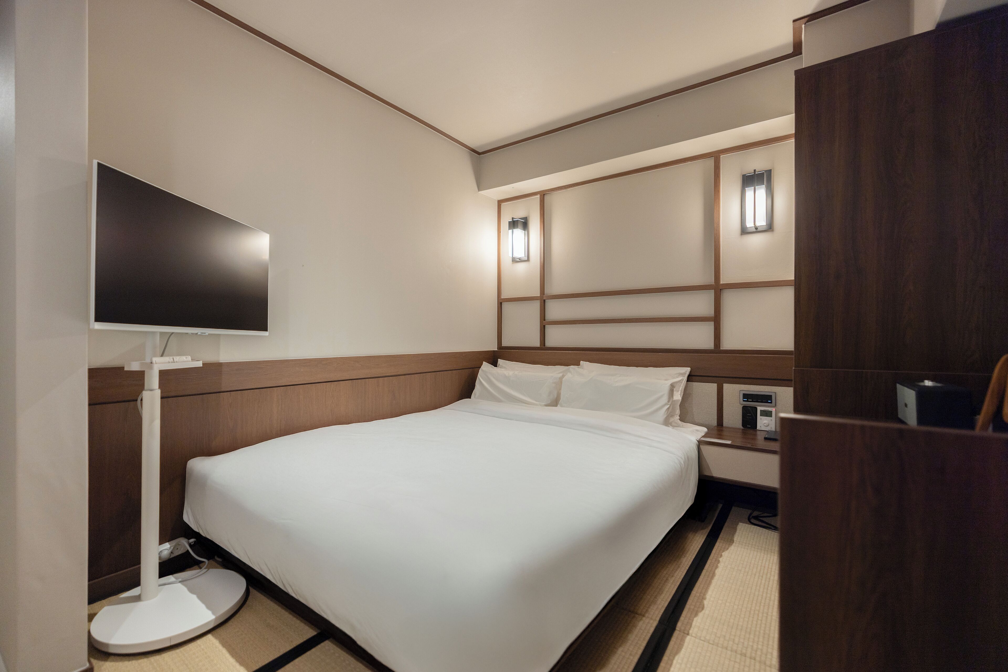 Habitación de hotel con cama, mesita de noche, televisor y luces fijadas en la pared.