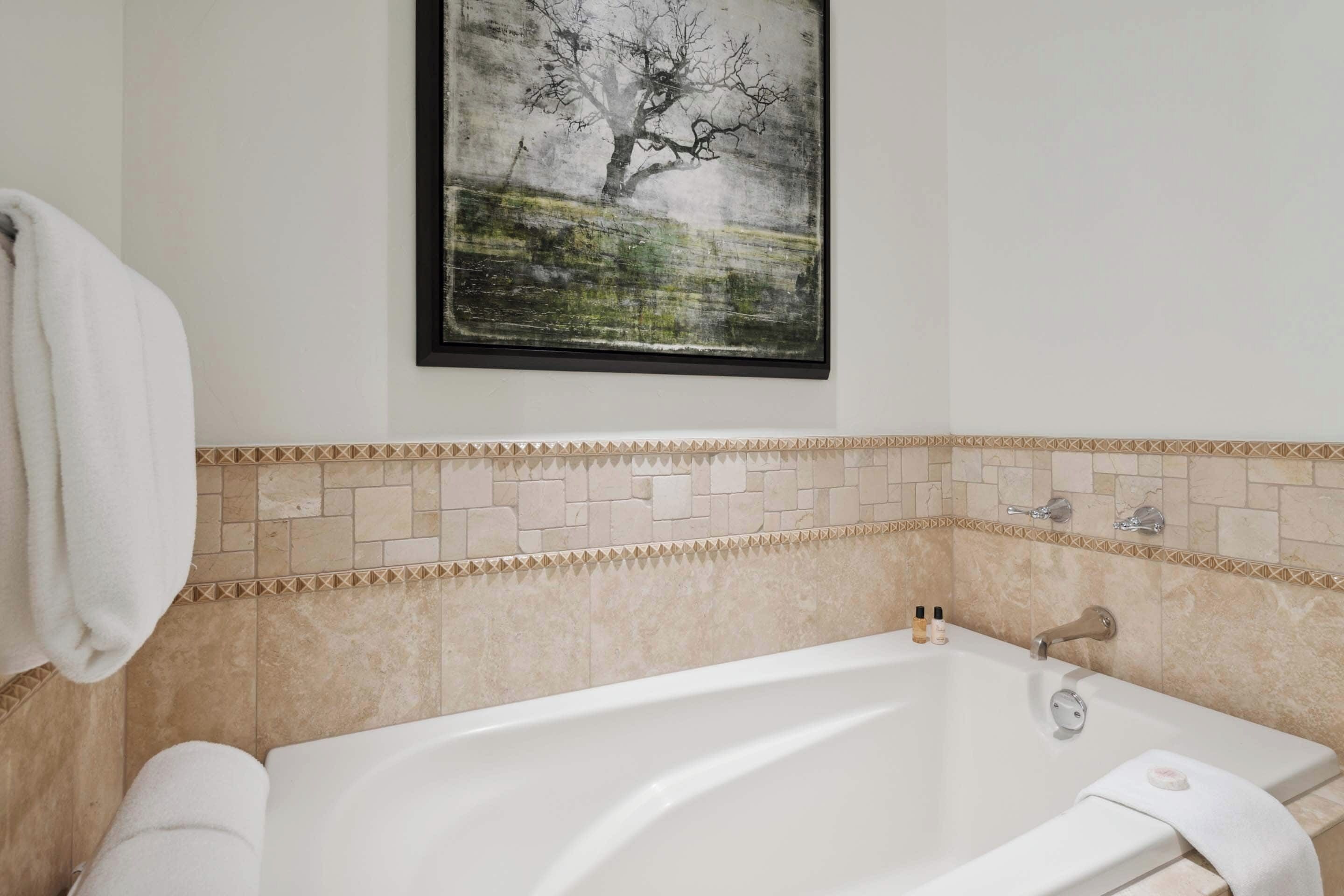 Une salle de bain moderne avec une baignoire sur pieds, des murs recouverts de carreaux beiges et une œuvre d’art encadrée au-dessus de la baignoire.