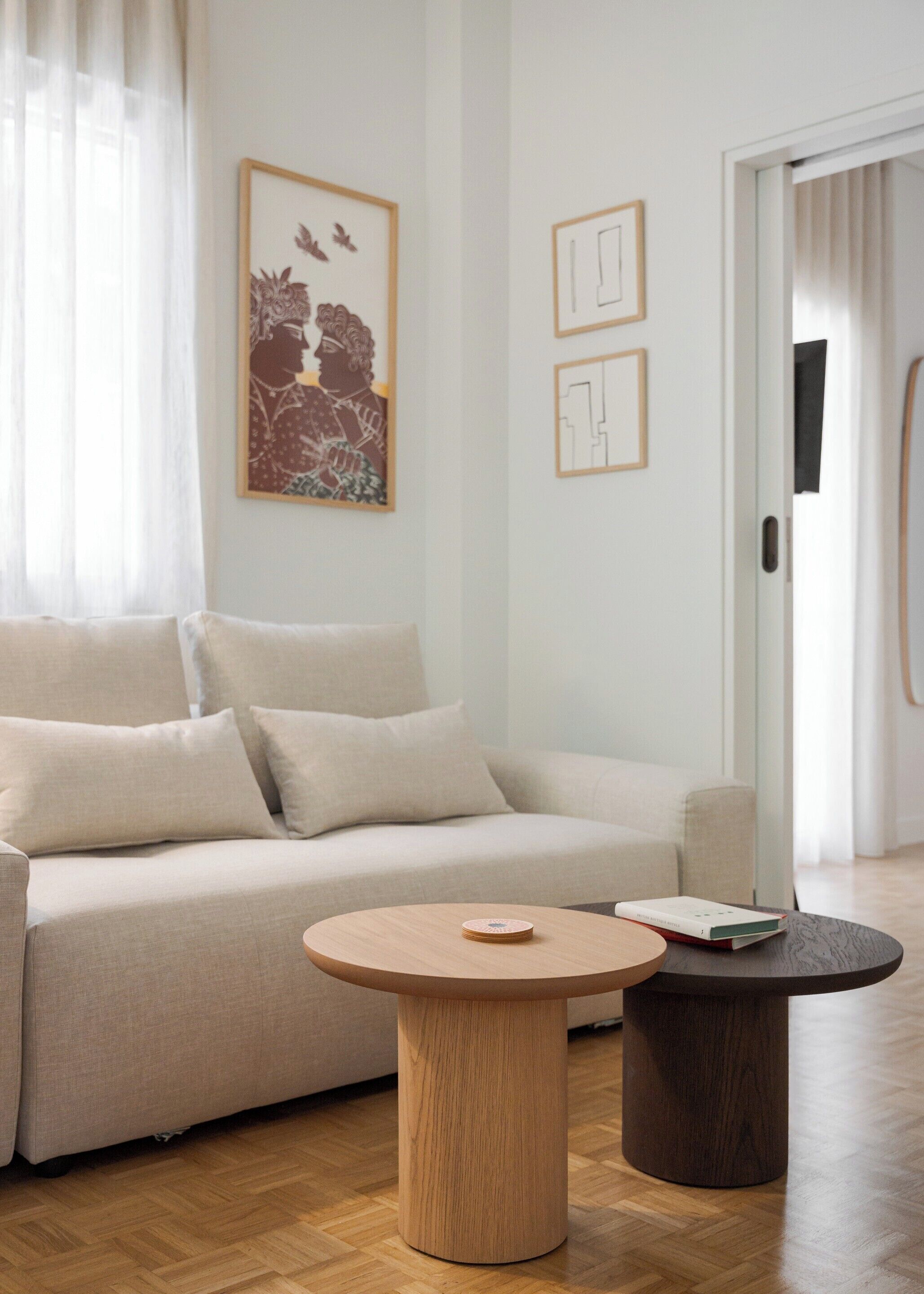 A modern living room with a beige sofa, two round wooden tables, and a framed artwork on the wall.