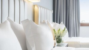 Ein ordentlich bezogenes Bett mit weißen Leinen und Kissen, ein Nachttisch mit einer Vase weißer Blumen und ein modernes Kopfstück mit vertikalen Linien.