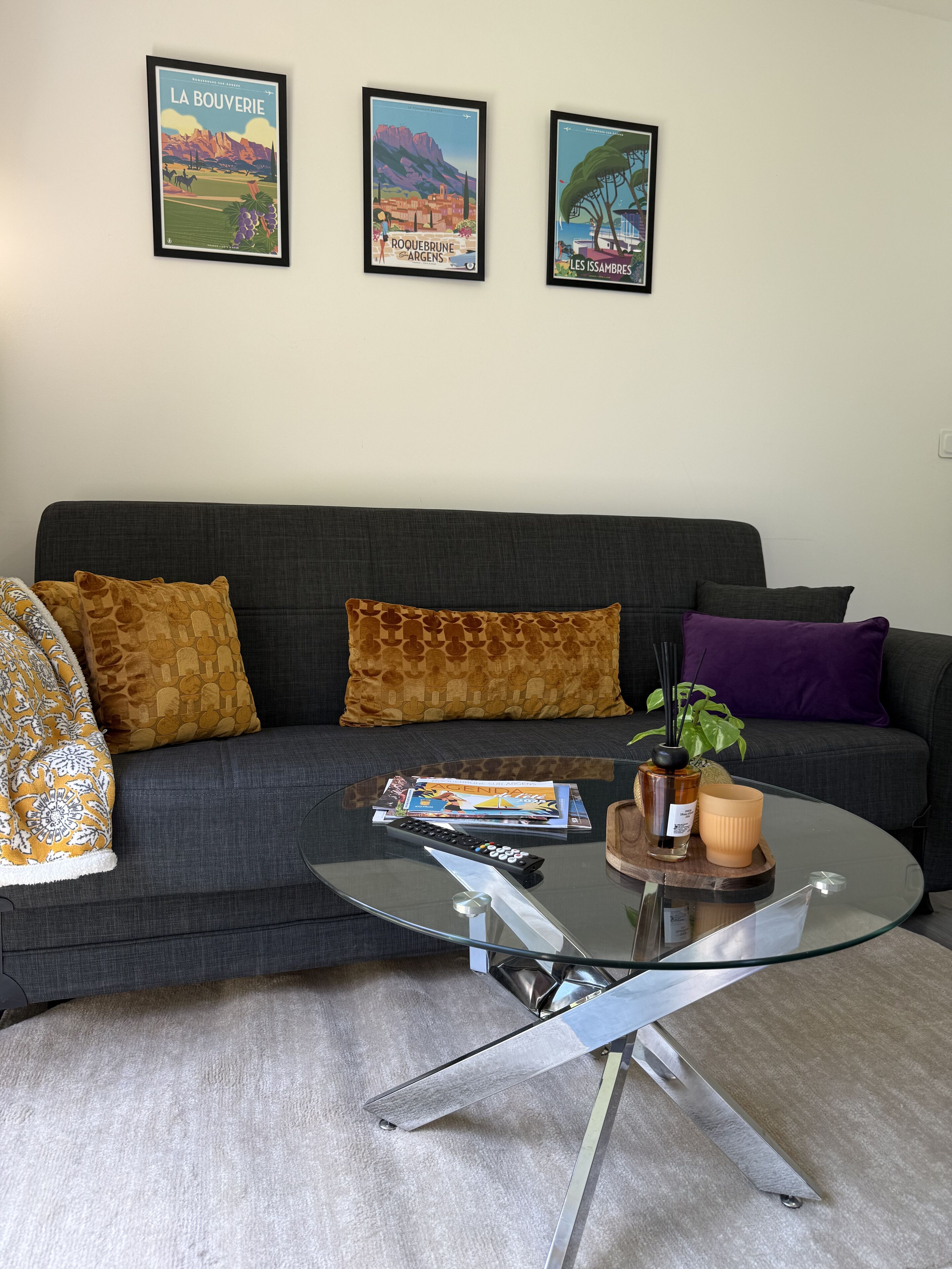 A modern living room with a dark grey sofa, patterned cushions, a glass coffee table with magazines and a remote control, and three framed posters on the wall.