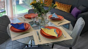 Comedor con mesa de vidrio preparada para cuatro personas, rodeada de sillas grises con tapicería. Un sofá con cojines estampados está apoyado en la pared, y un jarrón con flores amarillas sobre la mesa.