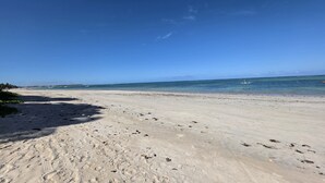 A wide sandy beach with footprints leading to the water, a clear blue sky, and a calm sea. - Nawa Beach Residence 301 by La Playa  (Tamandaré)