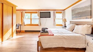 A bedroom with a bed, a TV mounted on the wall, a desk with a lamp, and a window with curtains.