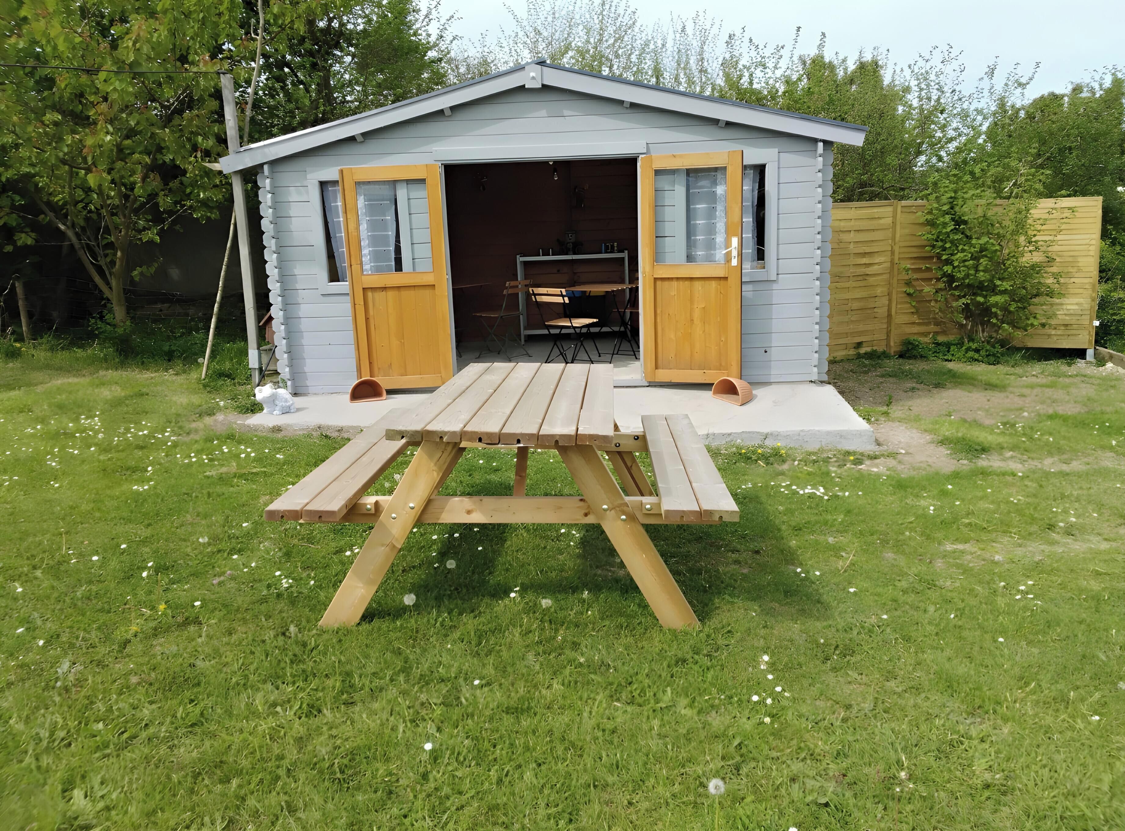 Een klein tuinhuisje met een openstaande deur, een picknicktafel ervoor en een houten hek op de achtergrond.