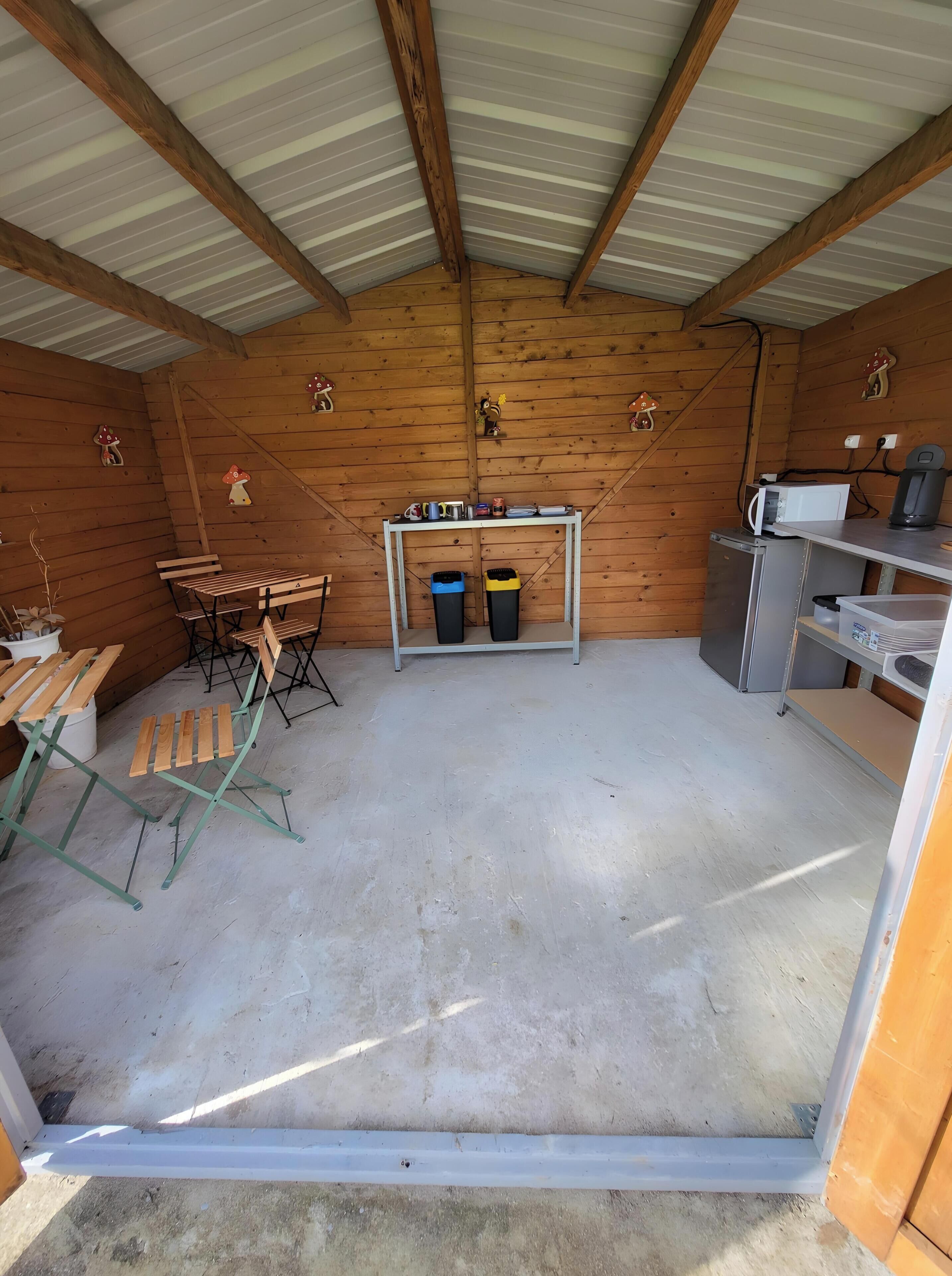 A small, enclosed space with wooden walls, a metal ceiling, a table with chairs, a trash bin, and a refrigerator.