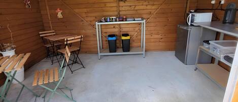 A small, enclosed space with wooden walls, a metal ceiling, a table with chairs, a trash bin, and a refrigerator.