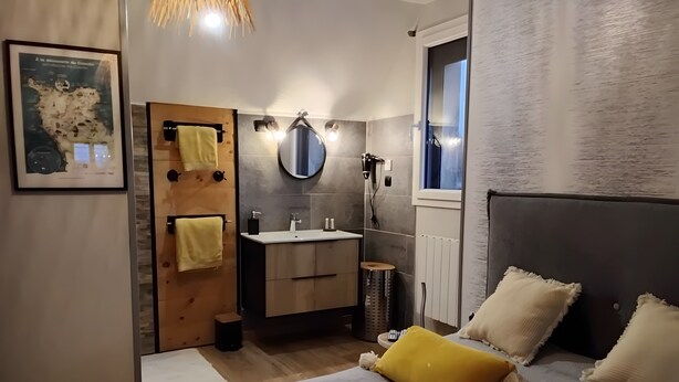 Een moderne slaapkamer met een bed, nachtkastje, spiegel, raam en een badkamer met wastafel en handdoekhouder.