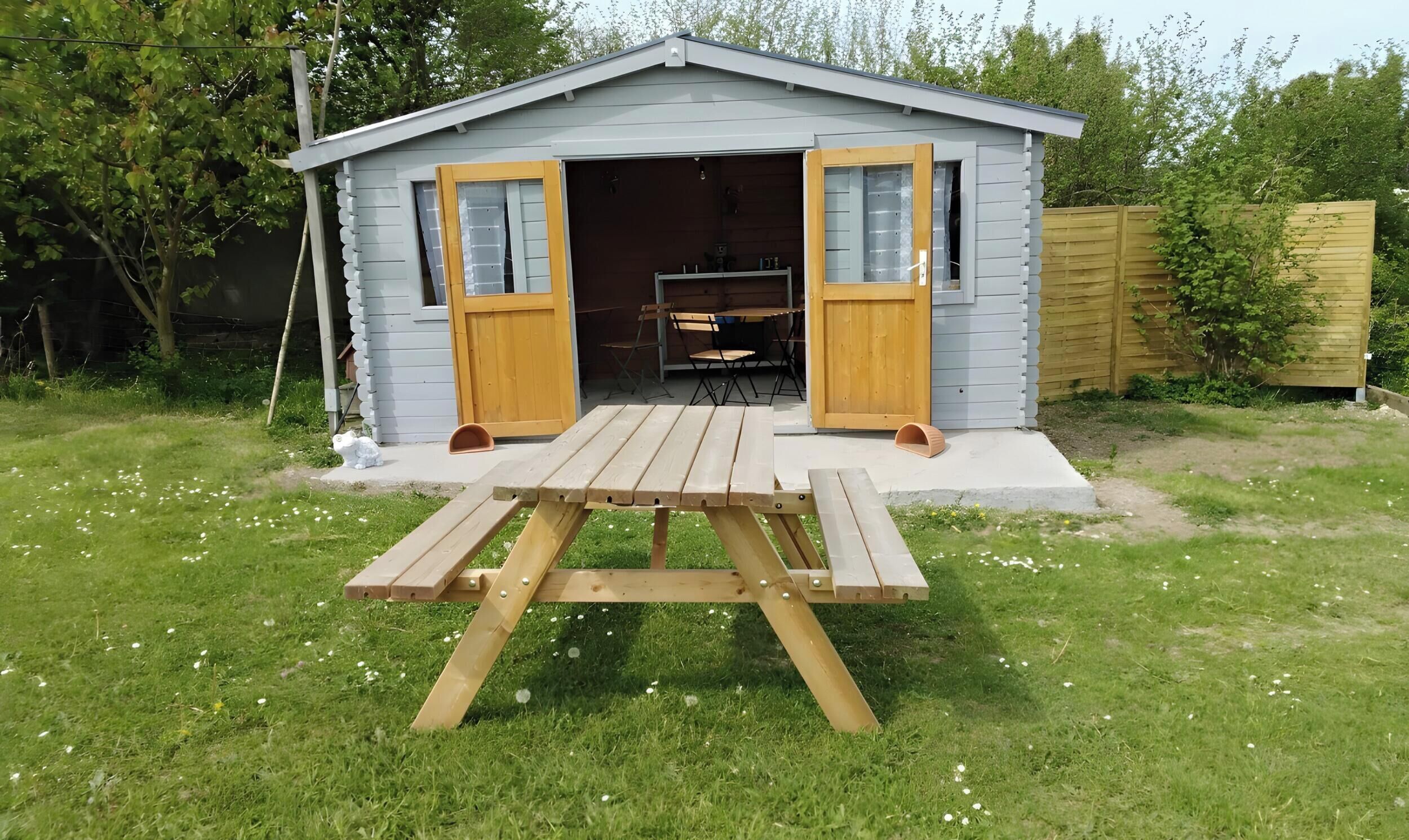 A small shed with two open doors, a picnic table in front, and a wooden fence in the background.