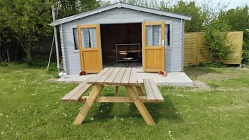 Un piccolo capanno con due porte aperte, un tavolo da picnic davanti e una recinzione in legno sullo sfondo.