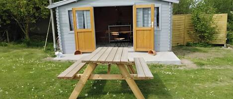 A small shed with two open doors, a picnic table in front, and a wooden fence in the background.