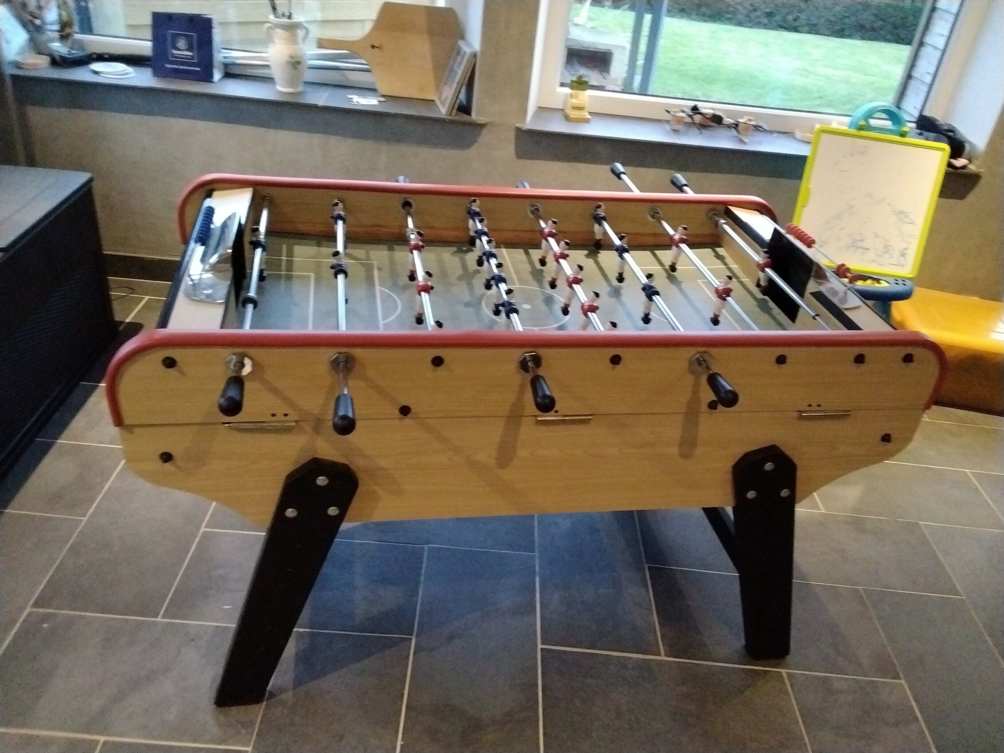 Een tafelvoetbaltafel in een kamer met betegelde vloer, een raam met uitzicht en een plank met diverse spullen.