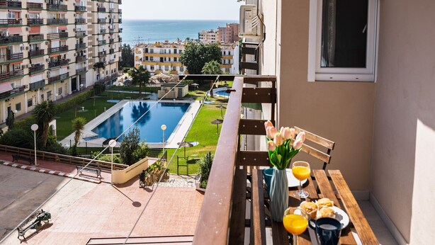 A balcony with a view of a swimming pool, a table with breakfast items, and a window overlooking the sea. - LAGOON VISTA BY ALFRESCO STAYS (Benalmádena)