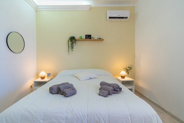 A neatly made bed with towels, bedside lamps, a wall-mounted air conditioner, and a shelf with decorative items.