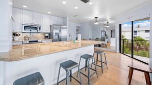 A modern kitchen with white cabinets, stainless steel appliances, a large island with bar stools, and a view of the outside through large windows. - Breathtaking Beach Views - 3bed on Siesta! (Siesta Key)
