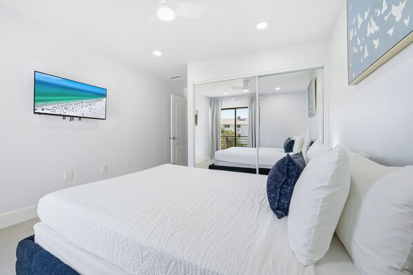 A modern hotel room with a large bed, a television displaying a beach scene, a mirror, and a view of the outdoors.