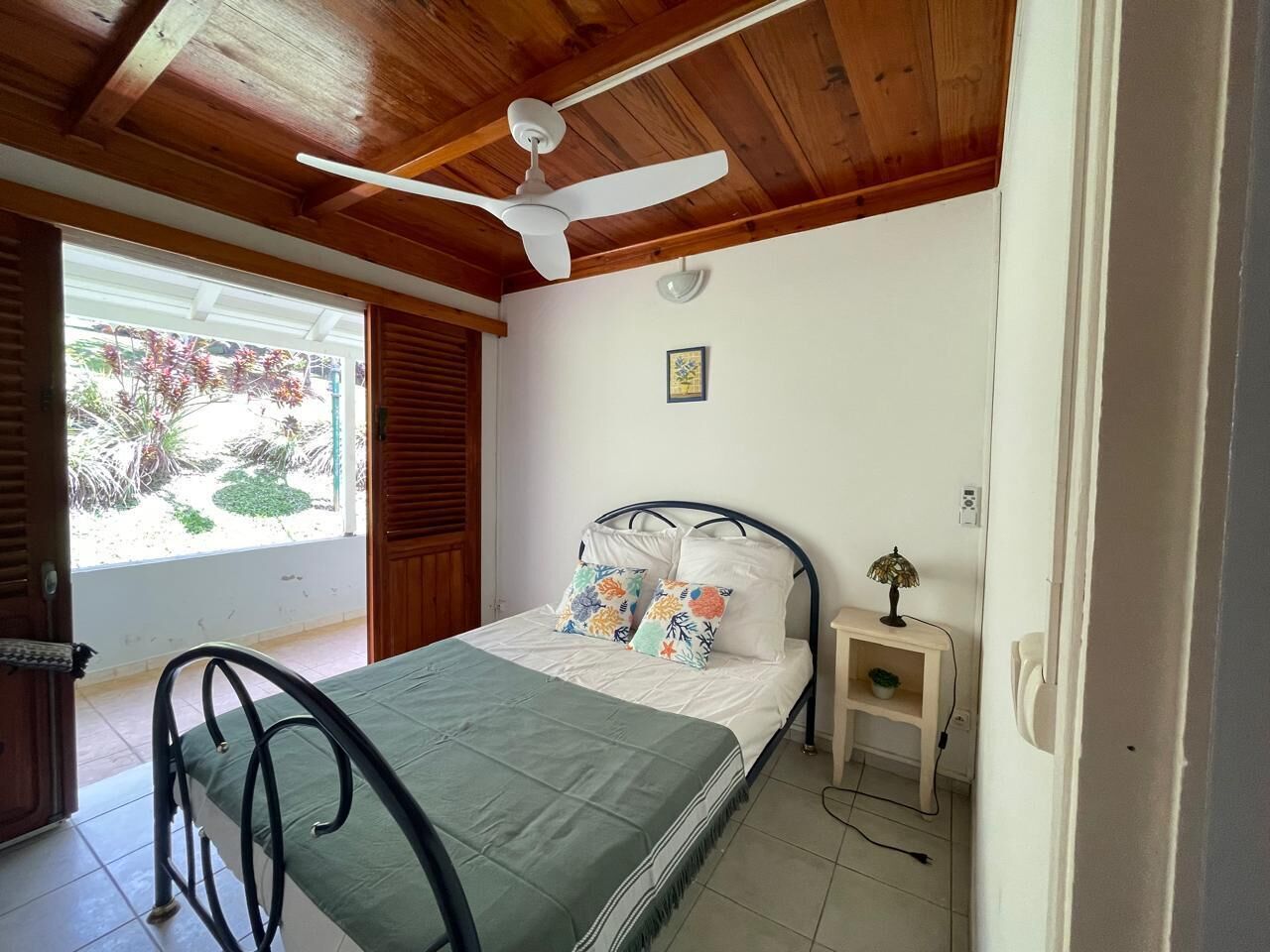 Een slaapkamer met een bed, nachtkastje, plafondventilator en een raam met uitzicht op groen.