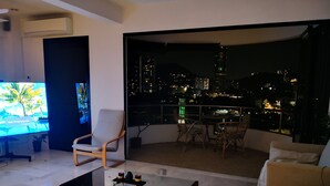 Un salon moderne doté d’un grand téléviseur, de sièges confortables, d’un balcon offrant une vue sur la ville et d’un coin repas.