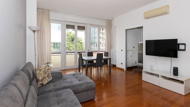 A modern living room with a grey sectional sofa, a dining table with chairs, a flat-screen TV, and a large window with curtains. - Large two-room apartment City Life - Mirable PM (Milano)