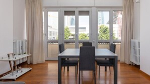 A modern dining room with a large table and four chairs, large windows with curtains, and a view of buildings and trees outside. - Large two-room apartment City Life - Mirable PM (Milano)