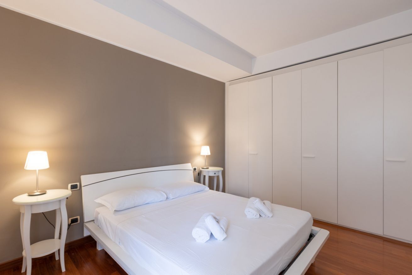 Een netjes opgemaakt bed met witte lakens en handdoeken, een nachtkastje met een lamp, en een inloopkast in een moderne hotelkamer.