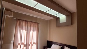 A hotel room with two beds, a ceiling light, a window with curtains, and a wall-mounted air conditioning unit. - Danis Hotel (Golem)