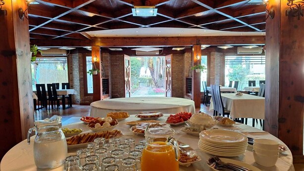 A breakfast buffet setup with various food items and beverages on a round table, surrounded by chairs and tables in a wooden interior. - Danis Hotel (Golem)