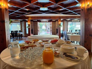 A breakfast buffet setup with various food items and beverages on a round table, surrounded by chairs and tables in a wooden interior. - Danis Hotel (Golem)