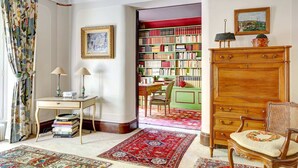 A room with a wooden cabinet, a chair, a desk with lamps, a bookshelf, and a patterned rug.